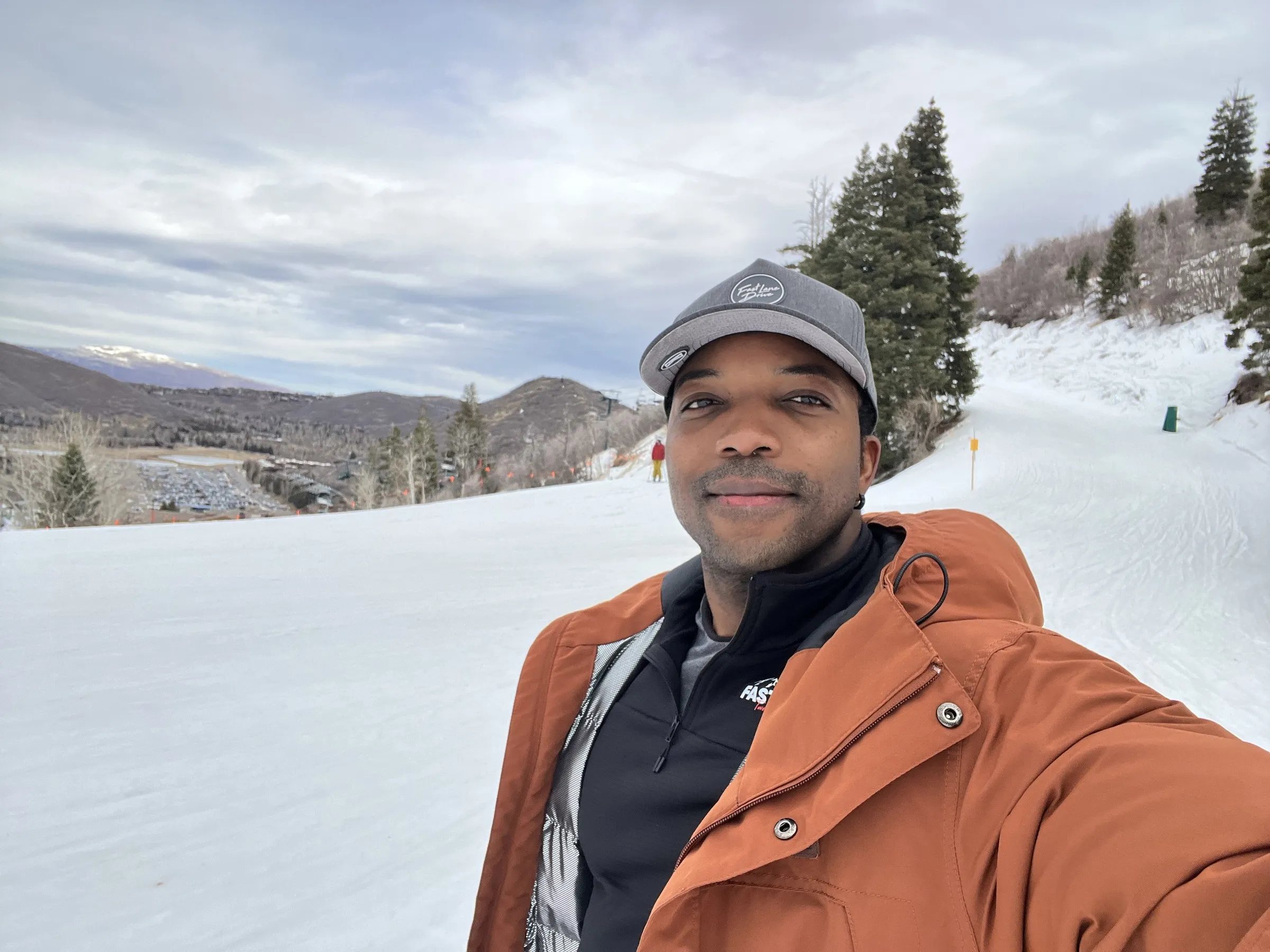 Mario G Bee on a ski slope in a grey cap and a rust jacket, mountains behind